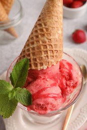 Photo of Delicious scoops of strawberry ice cream with mint and wafer cone in glass dessert bowl served on grey table, above view