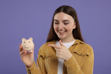 Happy woman pointing at piggy bank on purple background