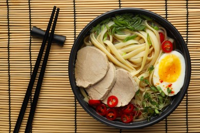 Photo of Delicious ramen with meat in bowl and chopsticks on bamboo mat, top view. Noodle soup