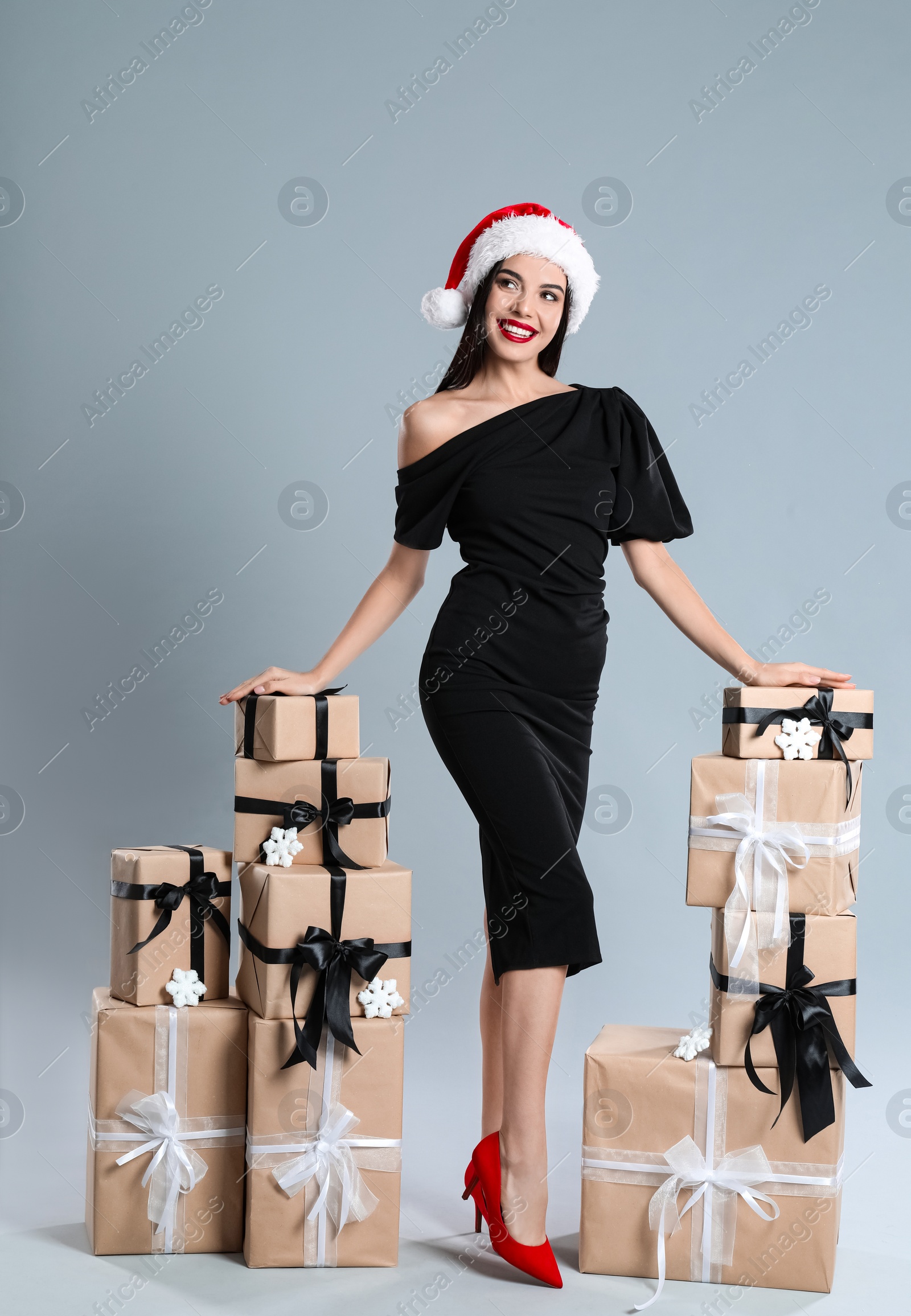 Photo of Woman in black dress and Santa hat with Christmas gifts on grey background