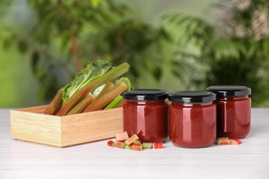 Photo of Jars of tasty rhubarb jam and stalks on white wooden table