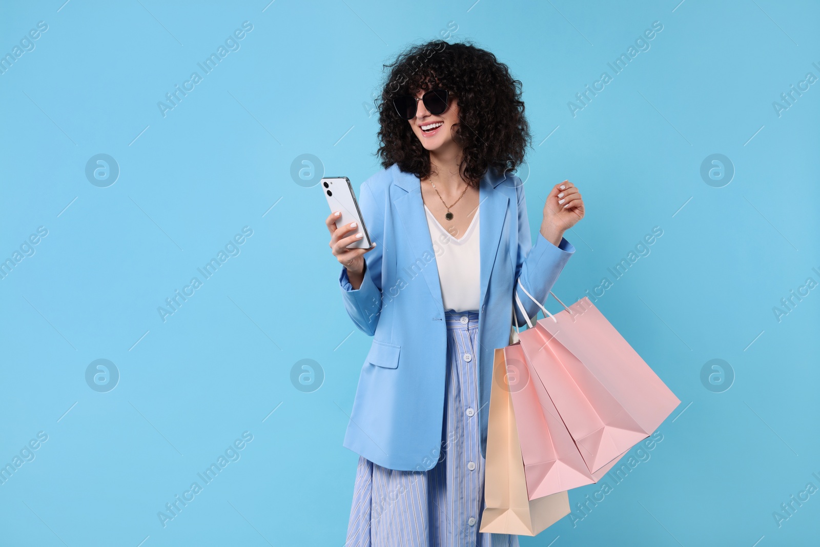 Photo of Happy young woman with shopping bags and modern smartphone on light blue background. Space for text
