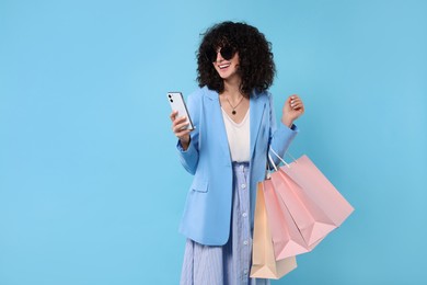 Happy young woman with shopping bags and modern smartphone on light blue background. Space for text