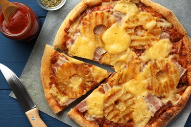 Delicious cut pineapple pizza, ingredients and knife on blue wooden table, flat lay
