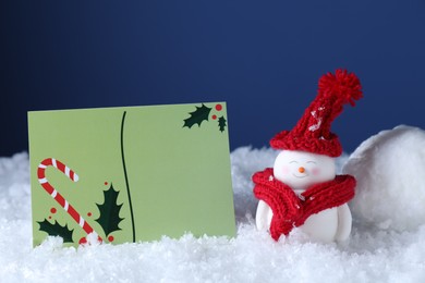 Photo of Cute decorative snowman and blank Christmas card on snow against blue background