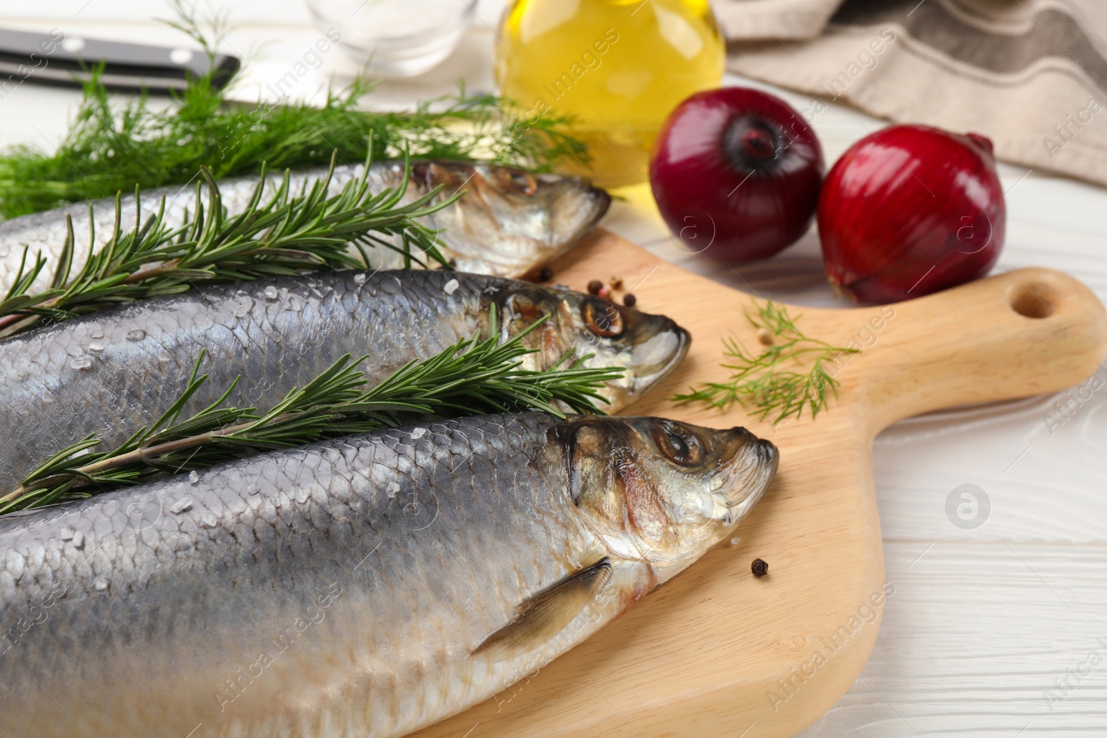 Photo of Board with delicious salted herrings, rosemary, onion and olive oil on white wooden table, closeup