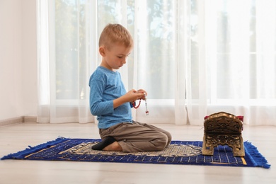 Little Muslim boy with misbaha and Koran praying on rug indoors