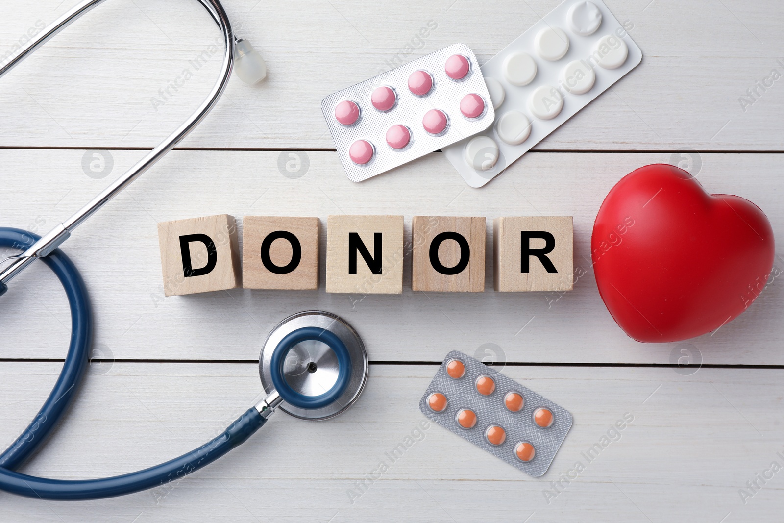 Photo of Word Donor made of cubes, stethoscope, pills and red heart on white wooden table, flat lay