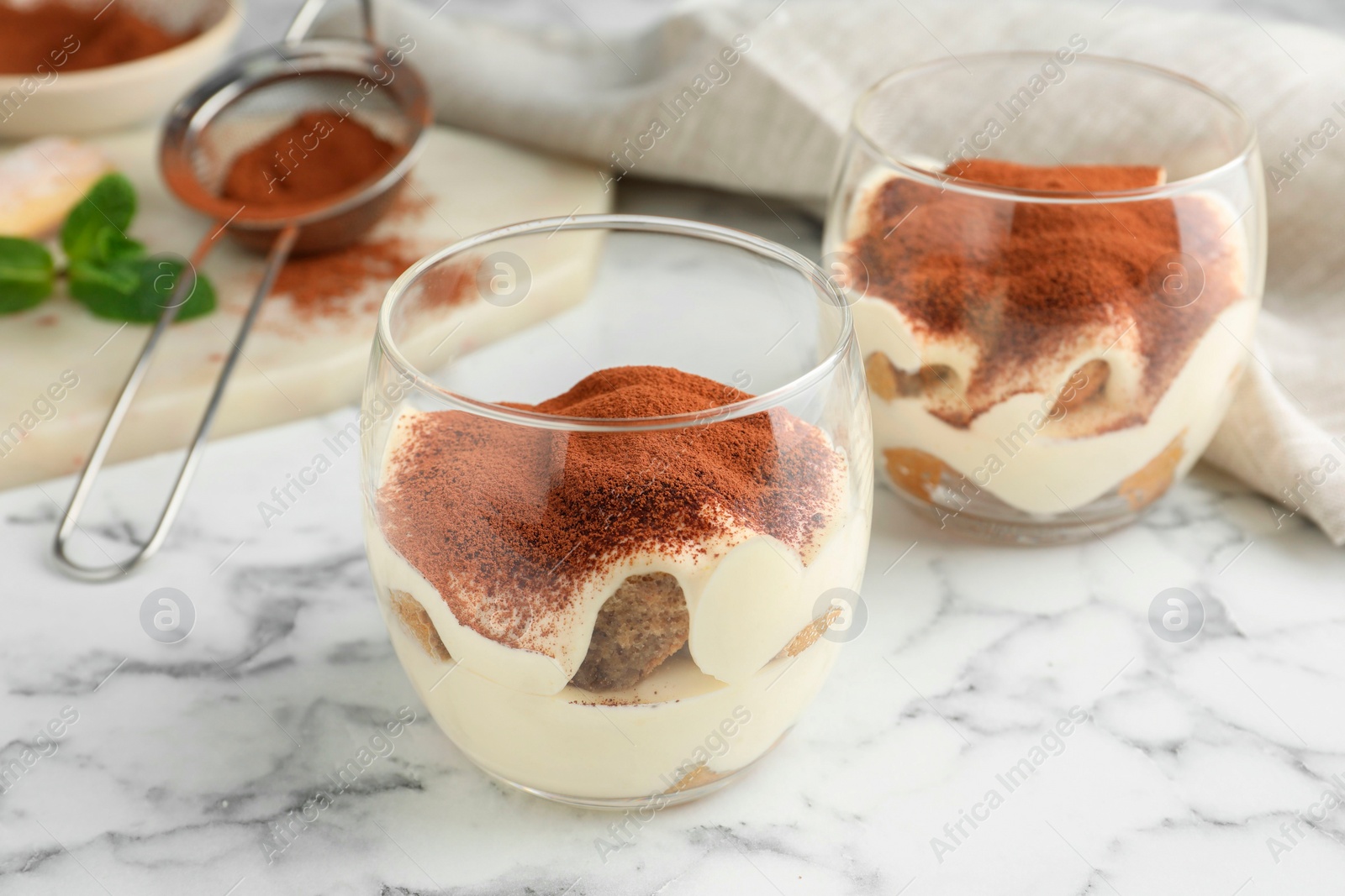 Photo of Delicious tiramisu in glasses and sieve with cocoa powder on white marble table, closeup