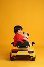 Little boy with his dog in toy car on yellow background, back view
