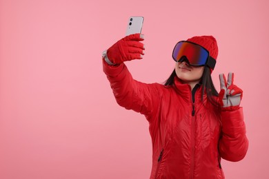 Woman in winter sportswear and goggles taking selfie and showing peace sign on pink background. Space for text