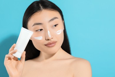 Photo of Beautiful young woman with sunscreen on her face holding sun protection cream against light blue background, space for text