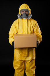 Photo of Man wearing chemical protective suit with cardboard box on black background. Prevention of virus spread