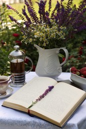 Open book, tea, ripe strawberries and bouquet of beautiful wildflowers on table in garden