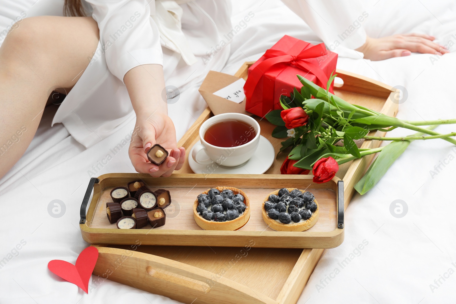 Photo of Tasty breakfast served in bed. Woman with desserts, tea, gift box and flowers at home, closeup