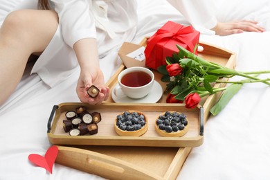 Photo of Tasty breakfast served in bed. Woman with desserts, tea, gift box and flowers at home, closeup