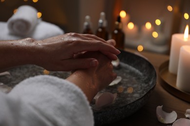 Photo of Woman soaking her hands in bowl of water and flower petals at table, closeup. Spa treatment