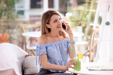 Young woman using mobile phone while drinking tasty healthy smoothie at table, indoors