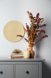 Photo of Stylish vases, dried eucalyptus branches and books on chest of drawers near white wall indoors. Interior design