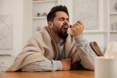Sick man wrapped in blanket with tissue at wooden table indoors. Cold symptoms