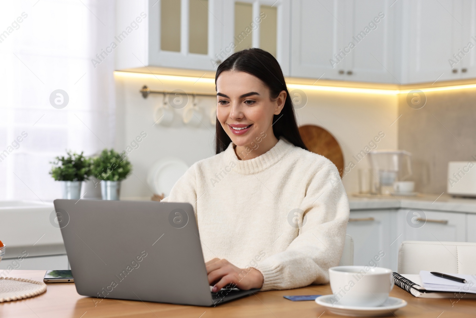 Photo of Happy young woman with laptop shopping online at wooden table in kitchen