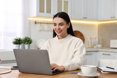 Photo of Happy young woman with laptop shopping online at wooden table in kitchen