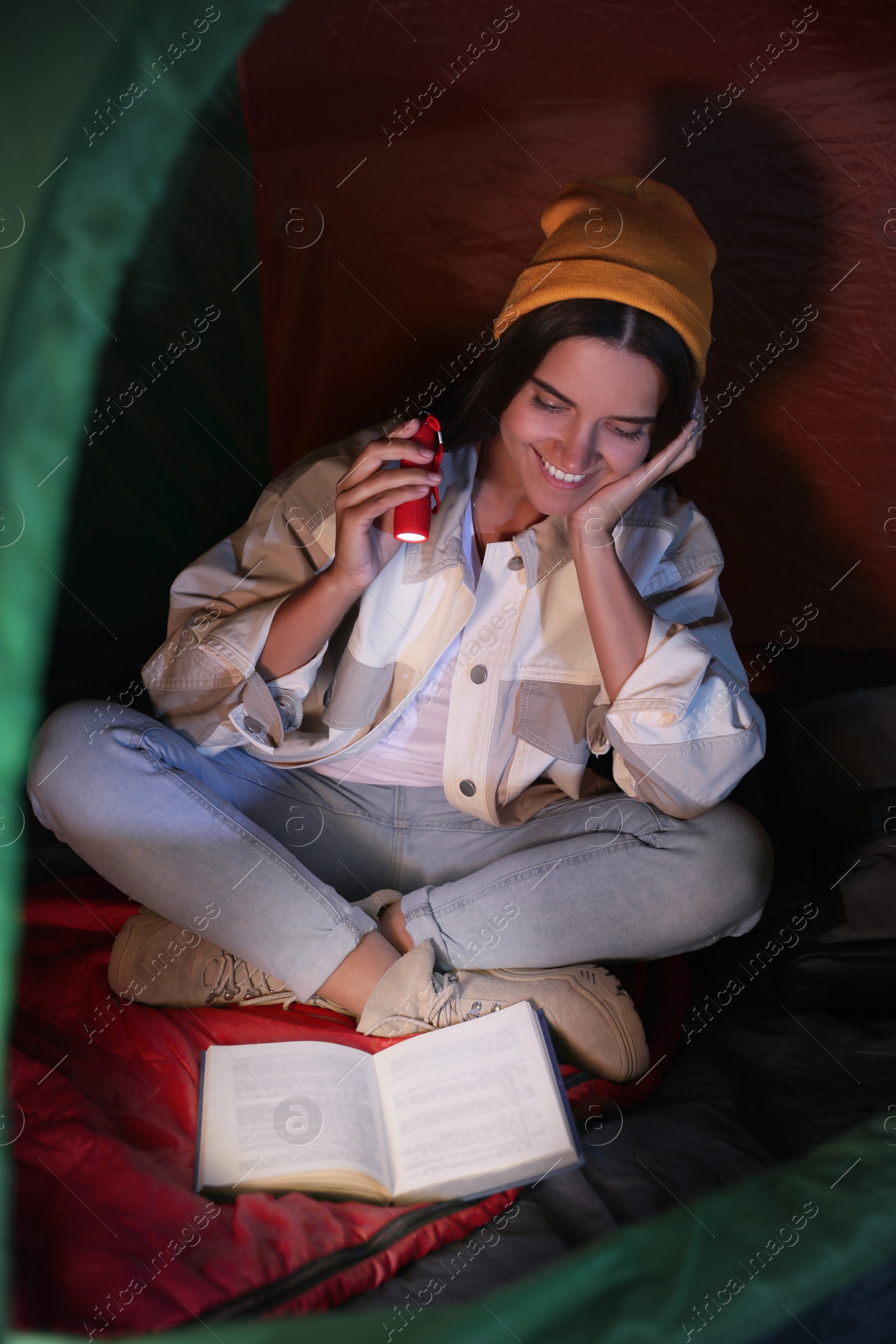 Photo of Young woman with flashlight reading book in tent at night