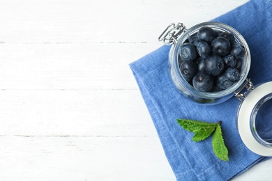 Photo of Glass jar of tasty fresh blueberries, mint leaves and fabric on white wooden table, flat lay with space for text