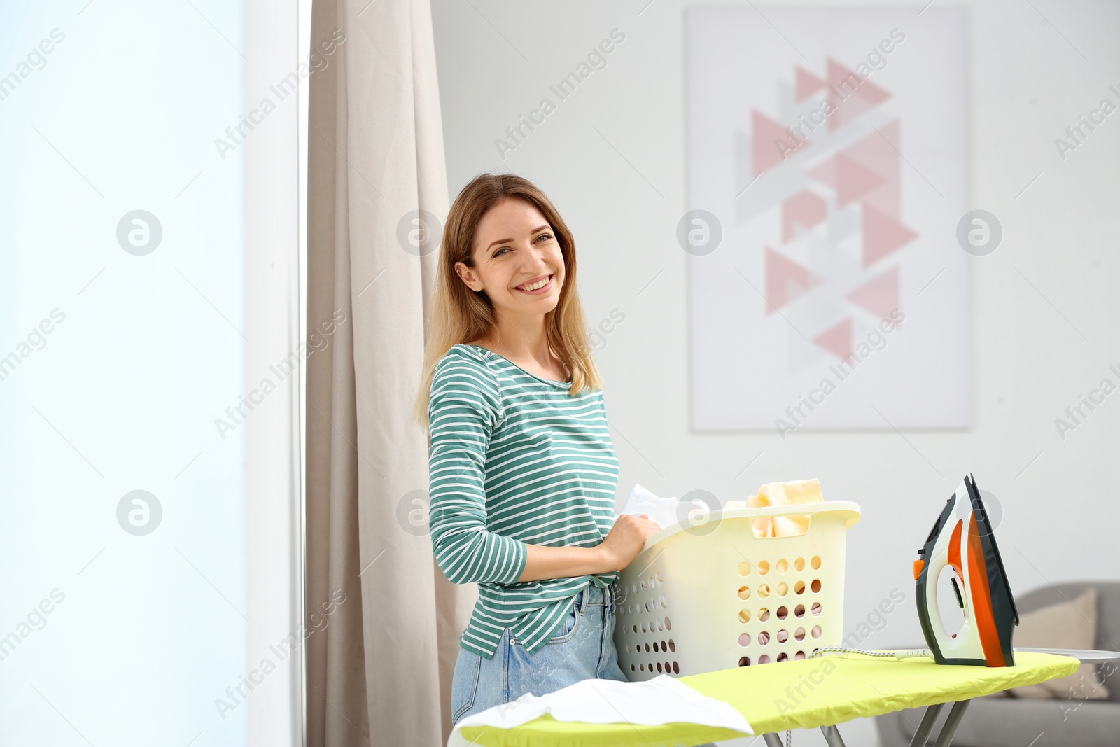 Photo of Young pretty woman holding basket of clean laundry at ironing board indoors