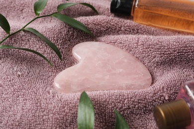 Rose quartz gua sha tool, green branches and cosmetic products on towel, closeup