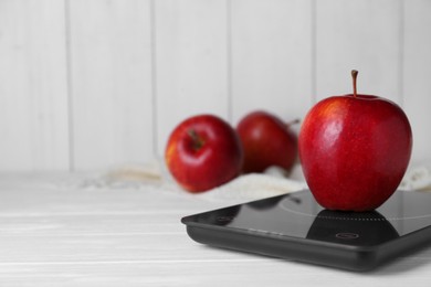 Photo of Digital kitchen scale with ripe red apple on white wooden table, space for text