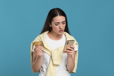 Photo of Confused woman with credit card and smartphone on light blue background. Debt problem