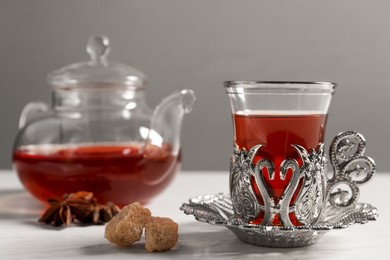 Photo of Glass of traditional Turkish tea in vintage holder, sugar and pot on white table, closeup