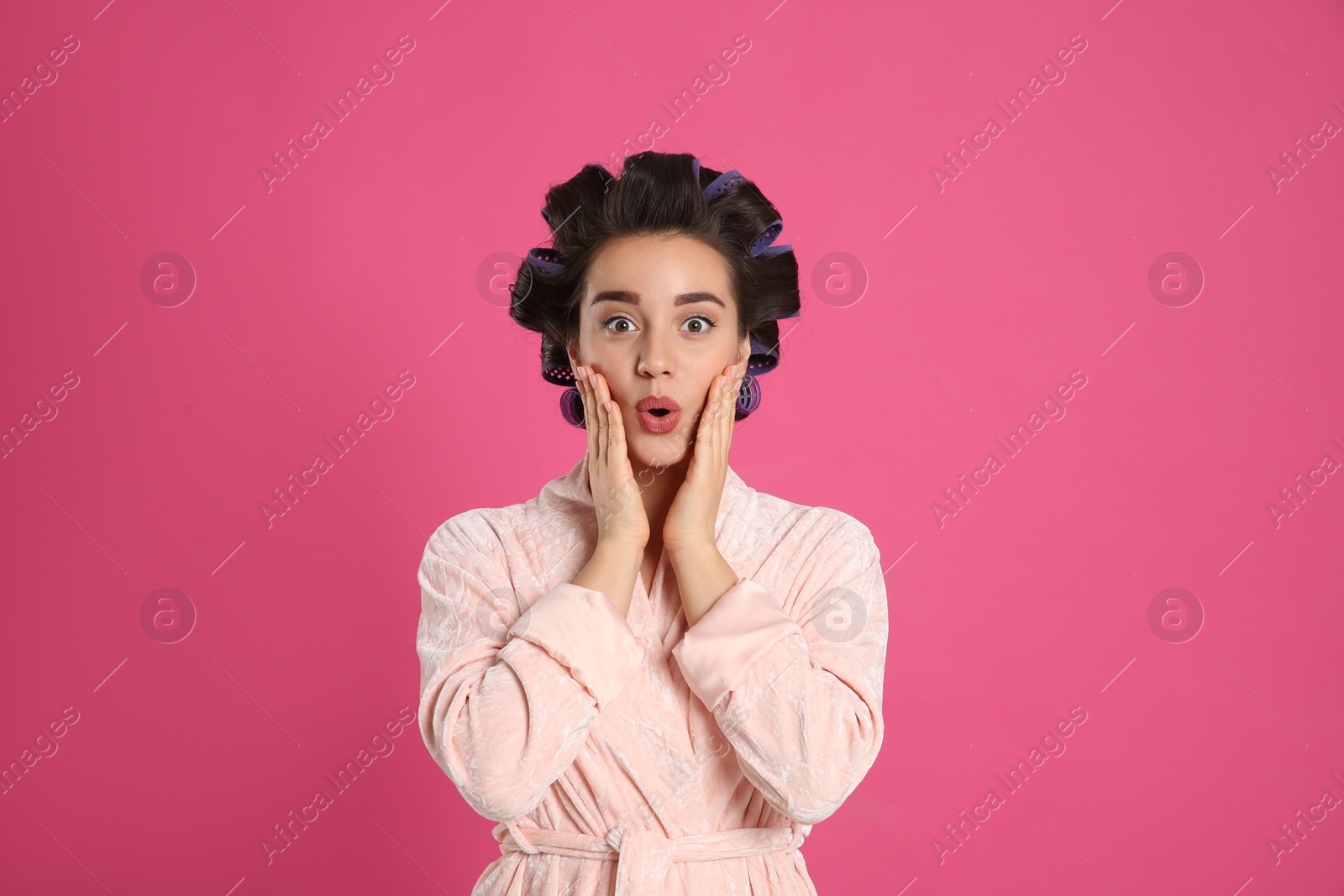 Photo of Emotional young woman in bathrobe with hair curlers on pink background