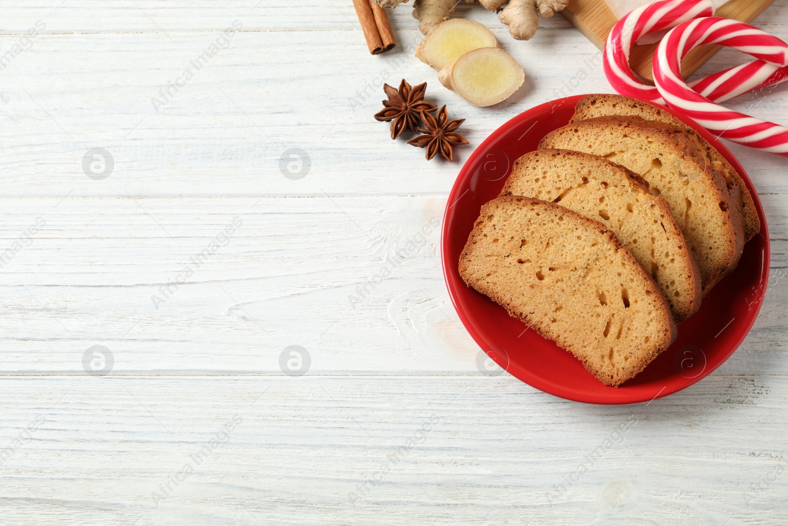 Photo of Slices of delicious gingerbread cake, ingredients and candy canes on white wooden table, flat lay. Space for text