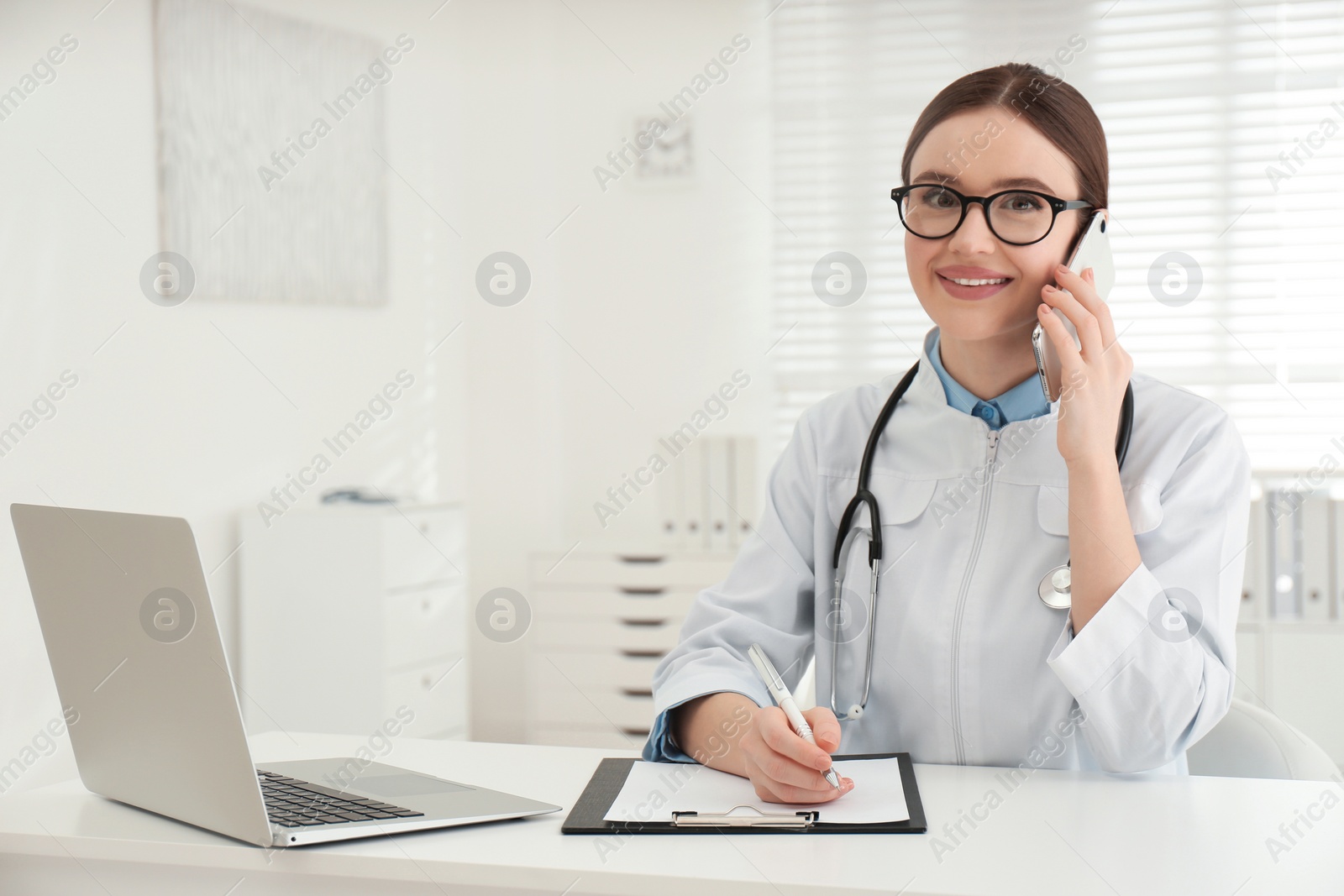 Photo of Young female doctor talking on phone at table in office