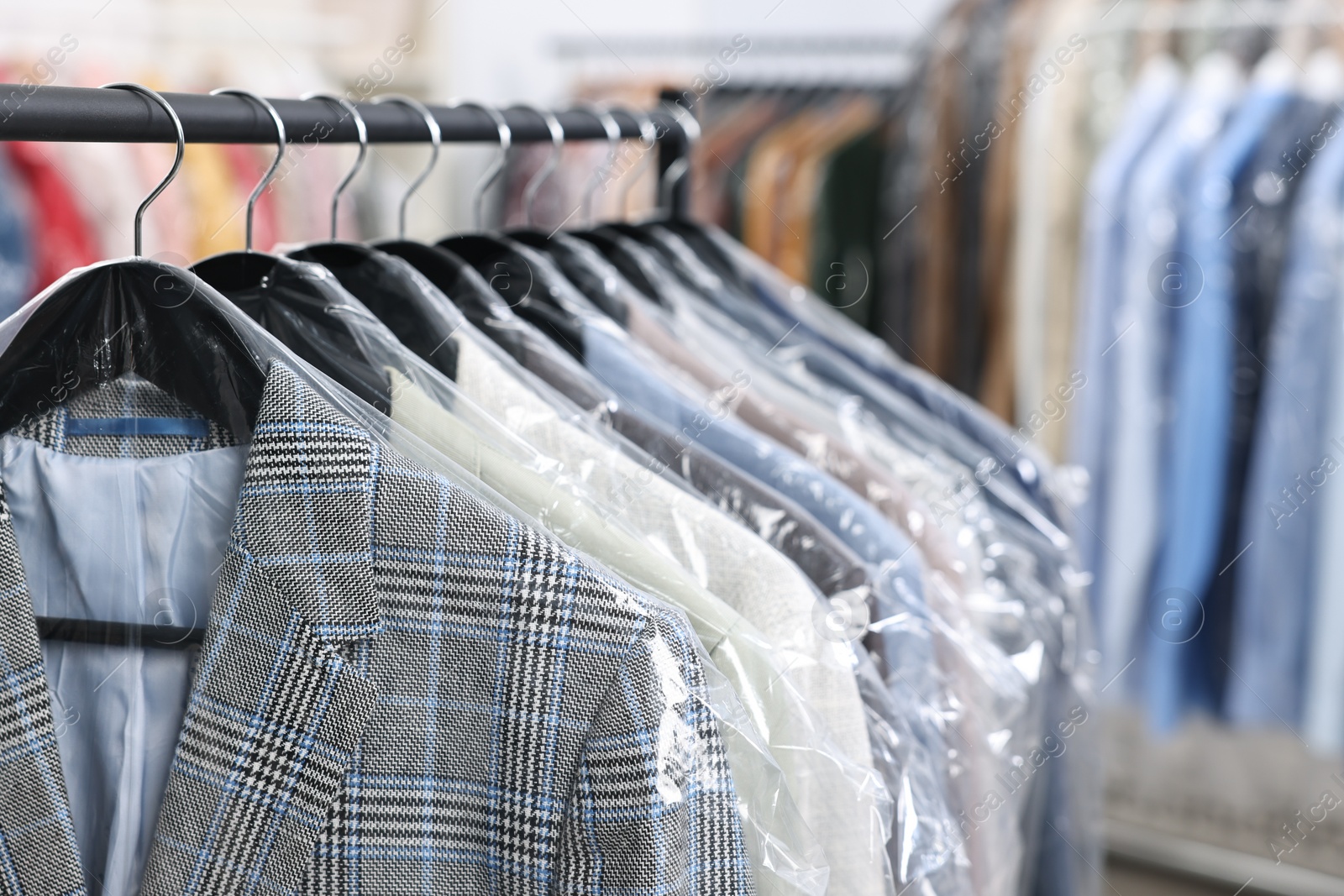 Photo of Dry-cleaning service. Many different clothes in plastic bags hanging on rack indoors, closeup