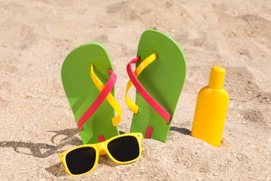 Photo of Stylish flip flops, sunglasses and sun protection cream on sand