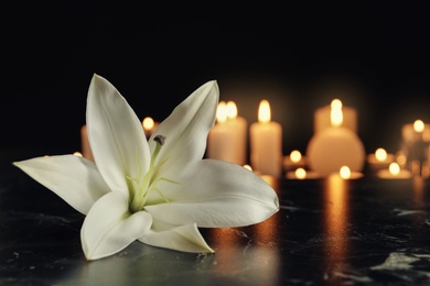 Photo of White lily and blurred burning candles on table in darkness, space for text. Funeral symbol
