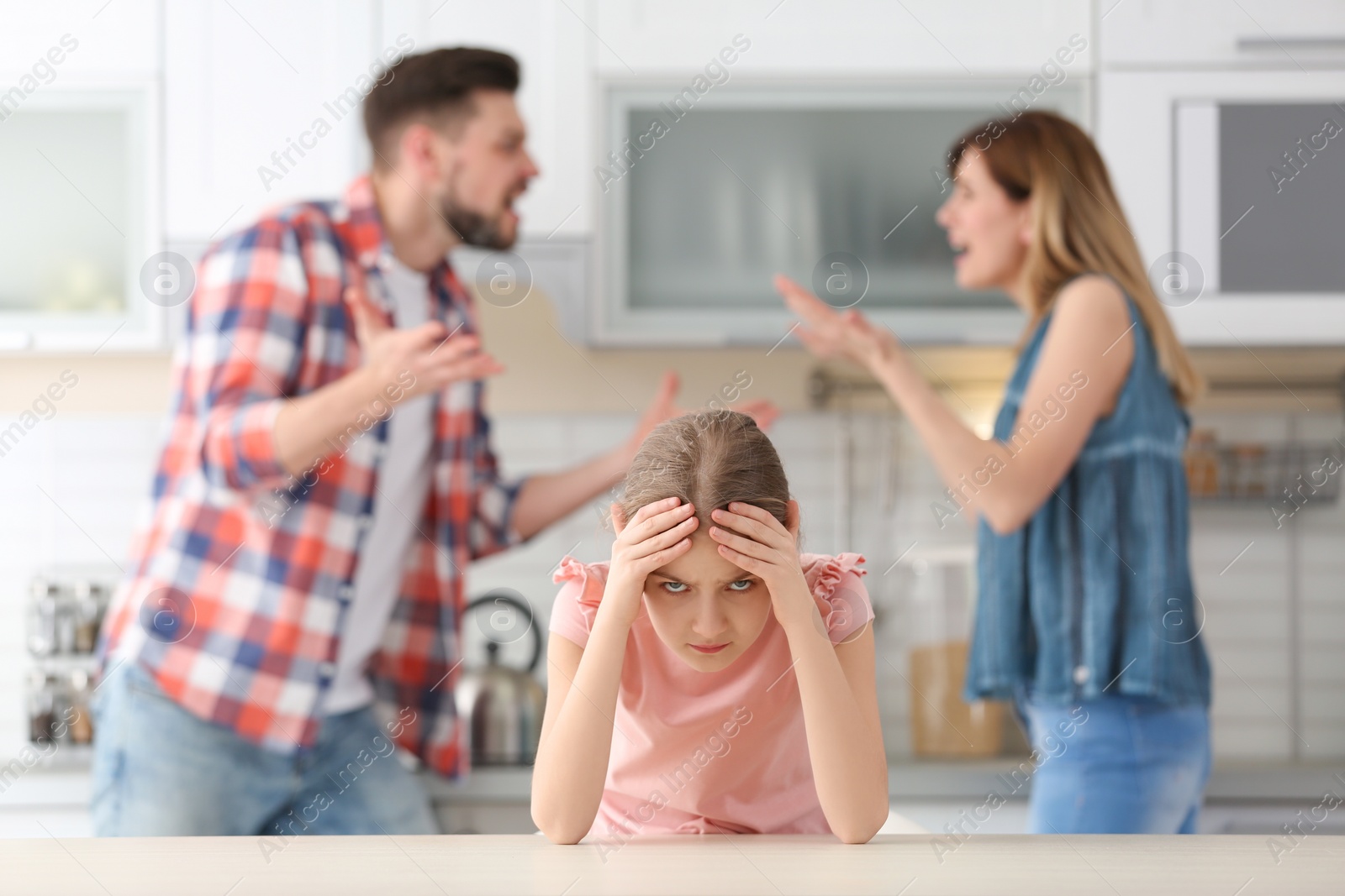 Photo of Little unhappy girl sitting at table while parents arguing on kitchen