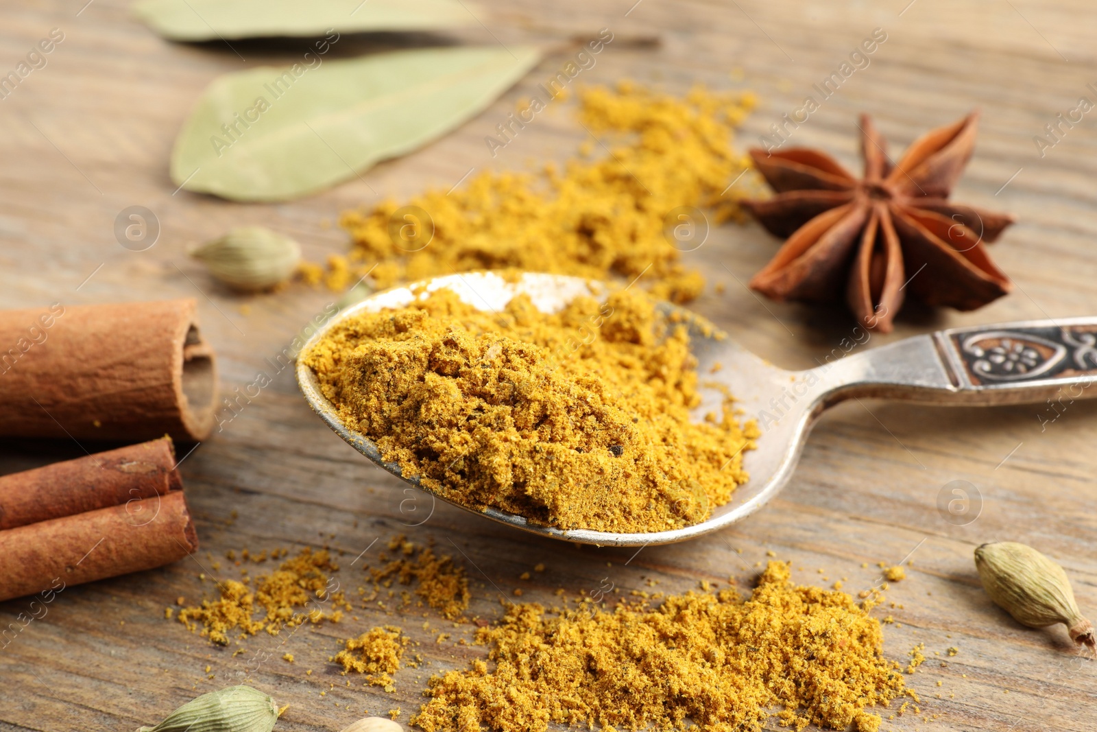 Photo of Spoon with dry curry powder and other spices on wooden table, closeup