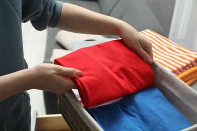 Photo of Woman putting folded clothes into storage basket indoors, closeup