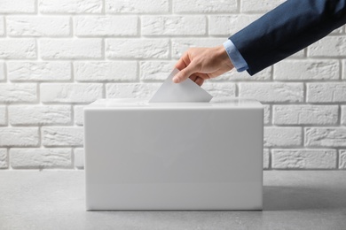 Man putting his vote into ballot box on table against brick wall, closeup