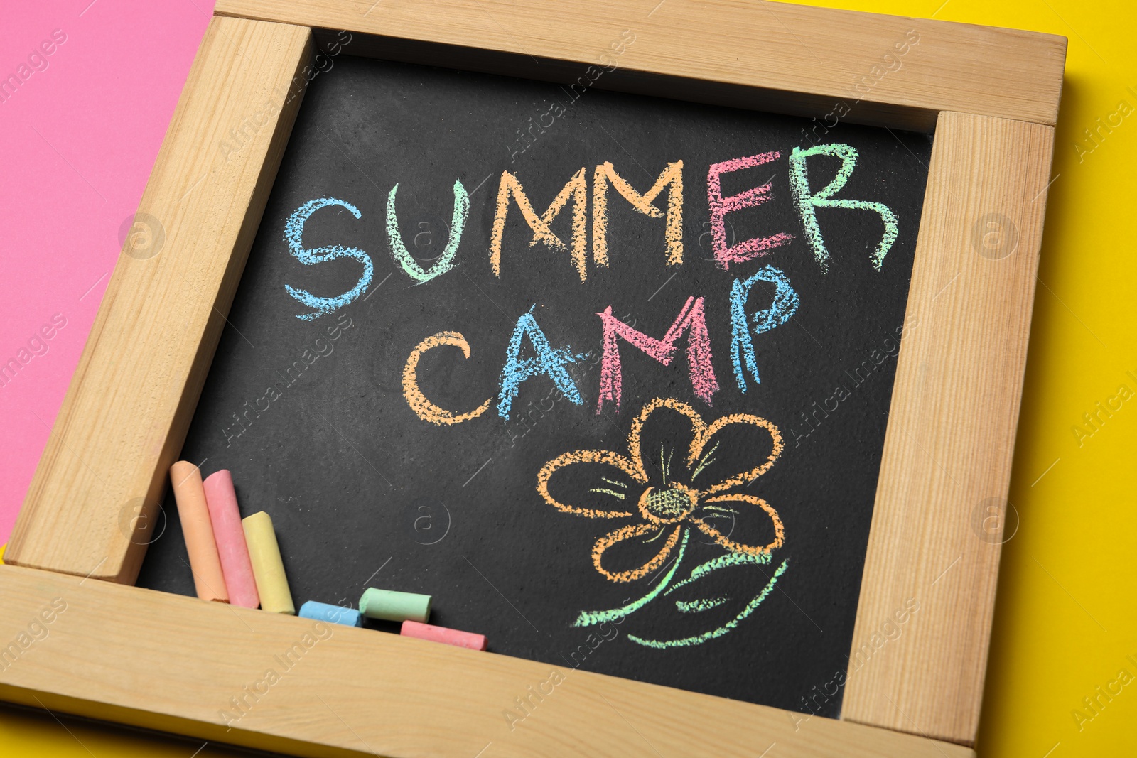 Photo of Small blackboard with text SUMMER CAMP, drawing and chalk sticks on color background