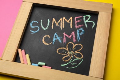 Photo of Small blackboard with text SUMMER CAMP, drawing and chalk sticks on color background