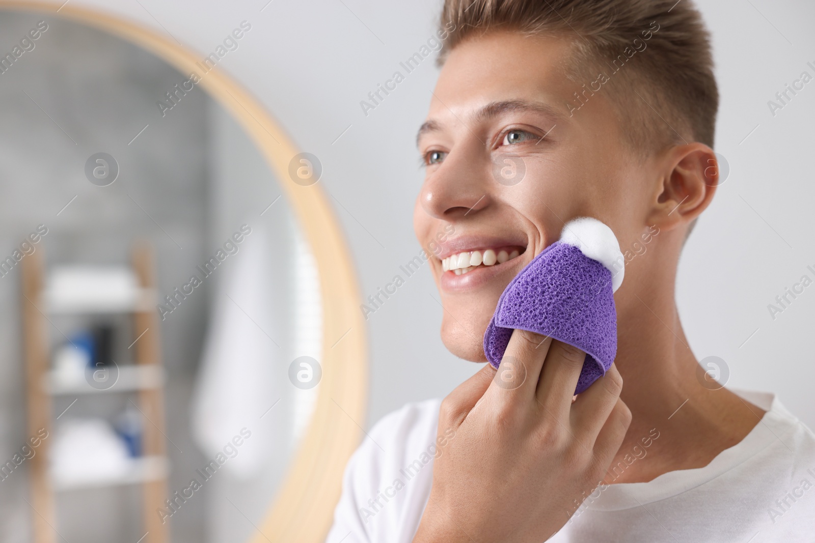 Photo of Happy young man washing his face with sponge in bathroom. Space for text