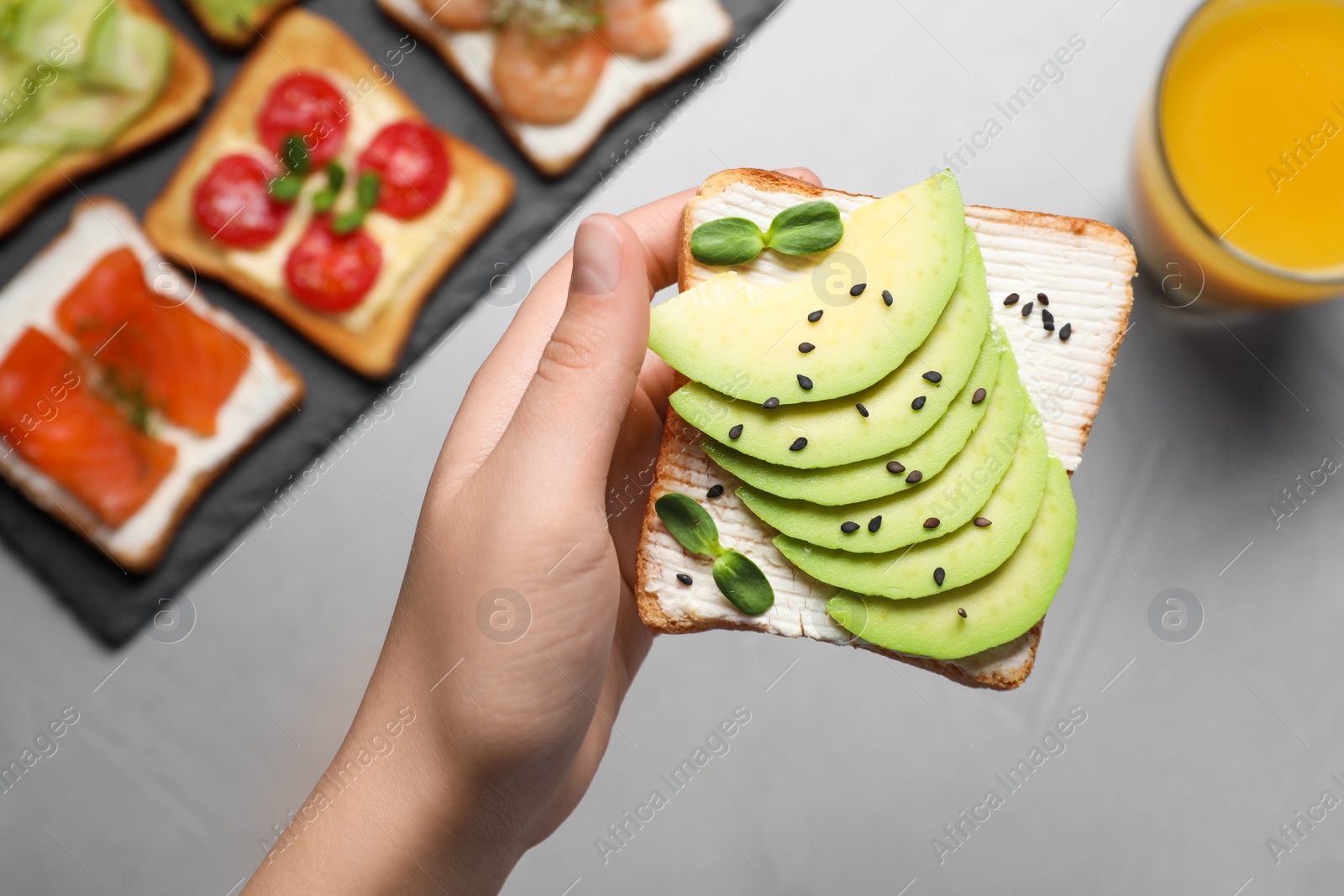 Photo of Woman holding delicious toast with cream cheese, avocado and sesame seeds at light grey table, top view