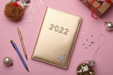 Photo of Golden planner with number 2022, stationery and festive decor on pink background, flat lay. New Year aims