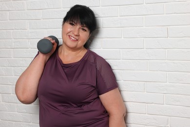 Happy overweight mature woman doing exercise with dumbbell near white brick wall. Space for text