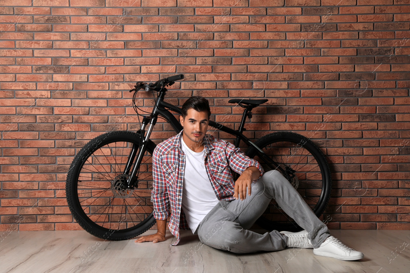 Photo of Handsome young man with modern bicycle near brick wall indoors